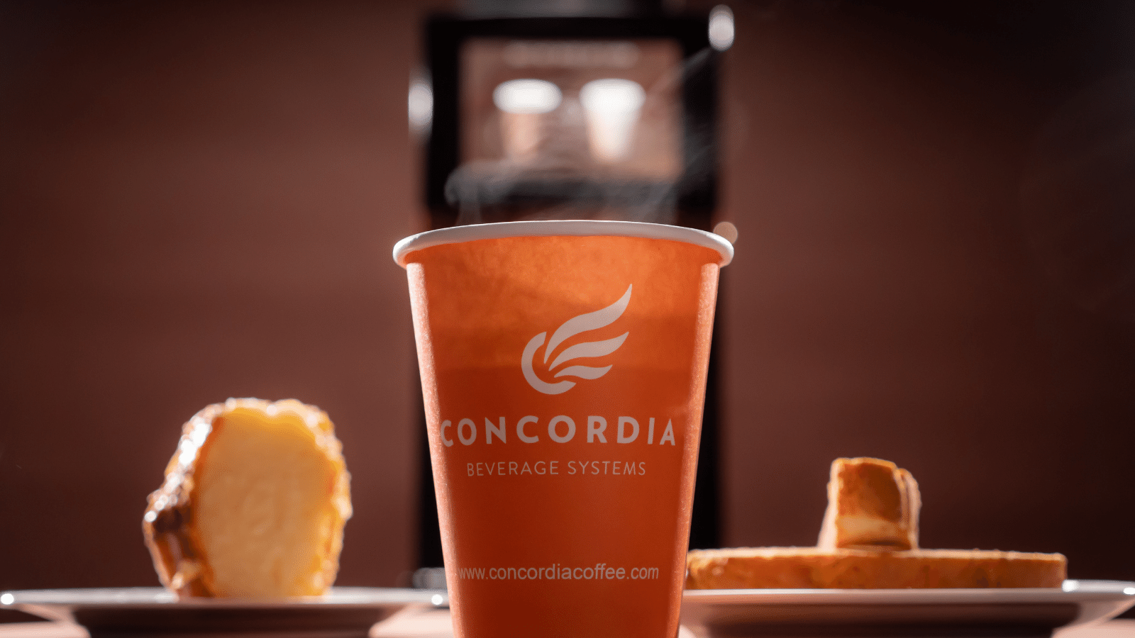 Coffee cup with Concordia logo on it in forefront. To left and right are breakfast breads. Behind blurred in background is a Concordia coffee maker.
